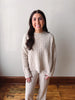 A woman wearing a matching cream cable-knit sweater and pants set, standing in front of a plain off-white wall with a wooden baseboard and hardwood floor.