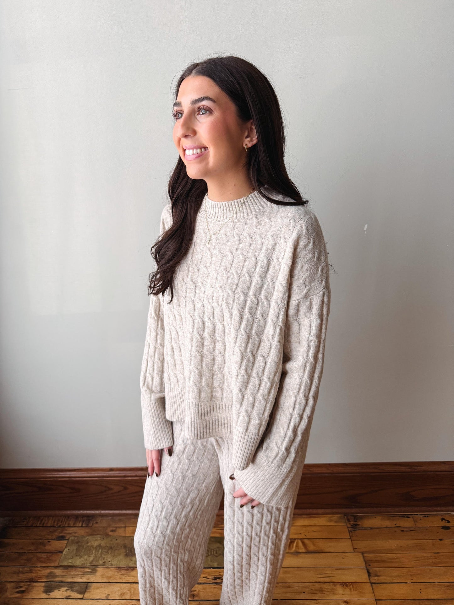 A woman wearing a matching cream cable-knit sweater and sweatpants set, standing in a room with a light gray wall and wooden floor.