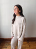 A woman wearing a matching cream cable-knit sweater and sweatpants set, standing in a room with a light gray wall and wooden floor.