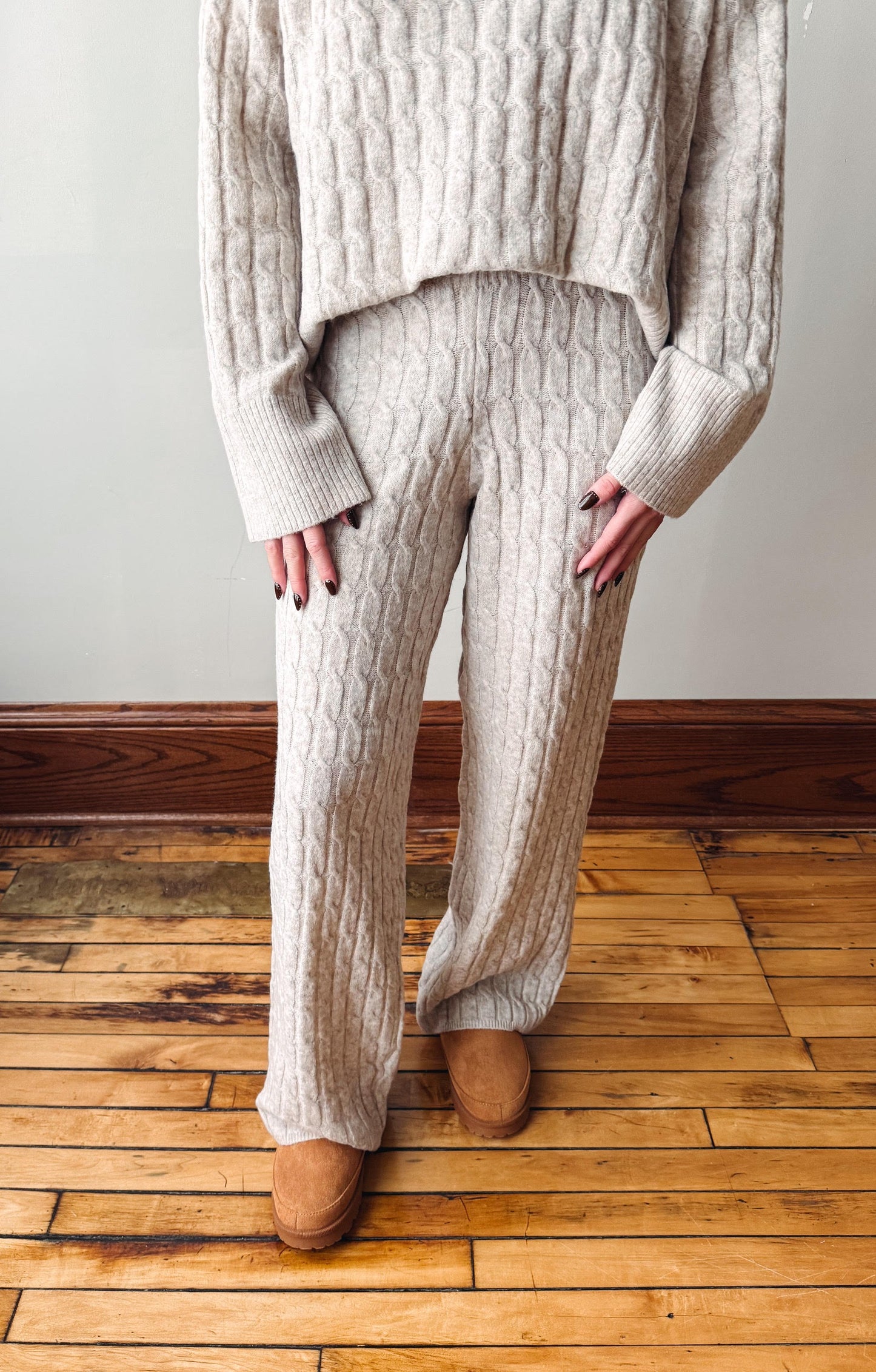 A person wearing a matching cream cable-knit sweater and wide-leg knit pants, standing on a wooden floor against a plain wall.