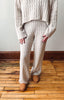 A person wearing a matching cream cable-knit sweater and wide-leg knit pants, standing on a wooden floor against a plain wall.