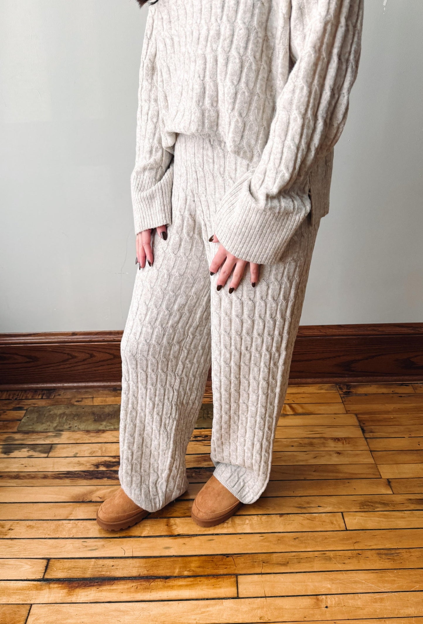A person wearing a cream cable-knit sweater and matching wide-leg pants, standing on a wooden floor against a light wall.