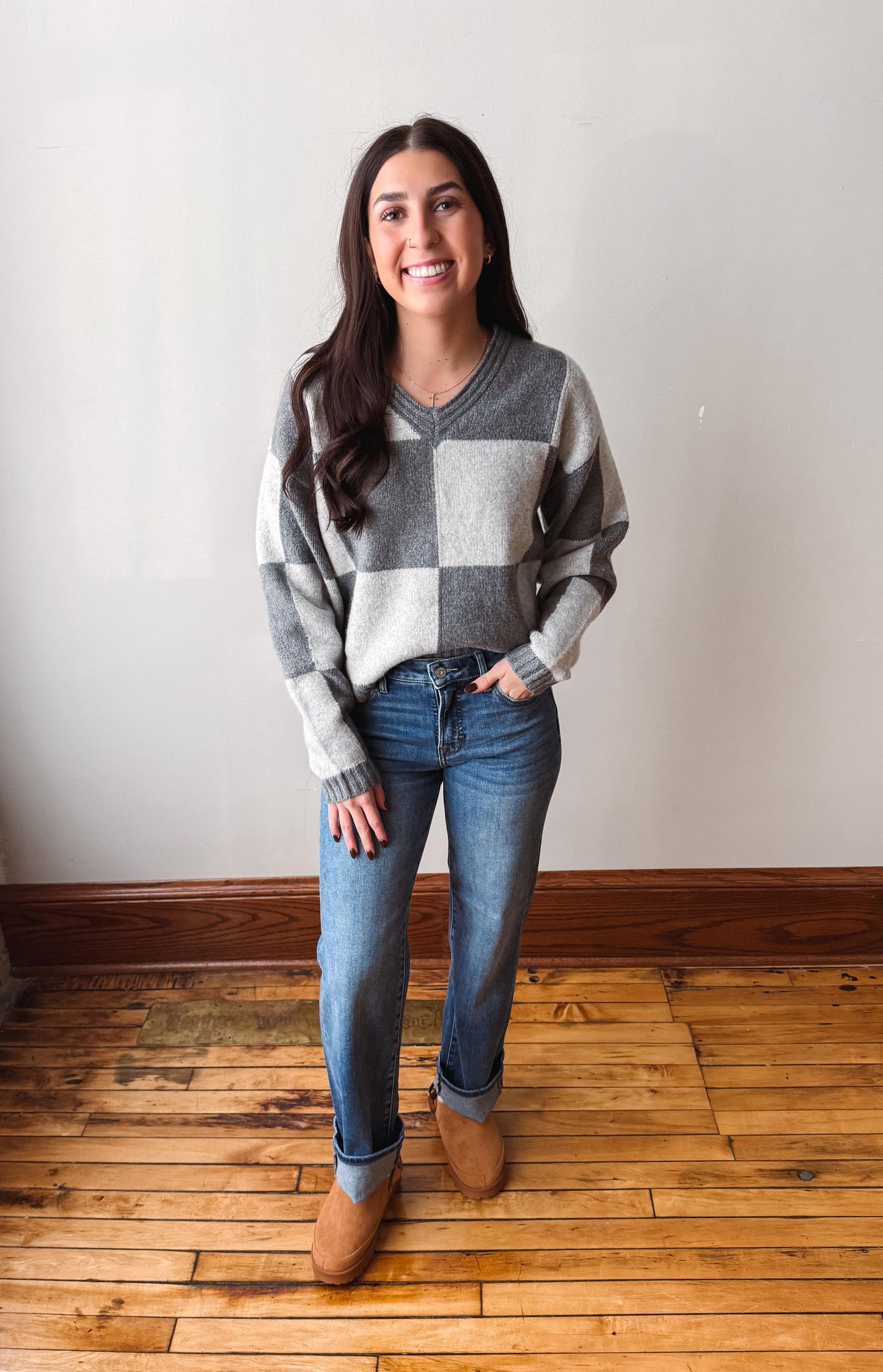 A woman wearing a gray and white checkered V-neck sweater, blue jeans, and brown suede boots, standing on a wooden floor against a plain off-white wall.