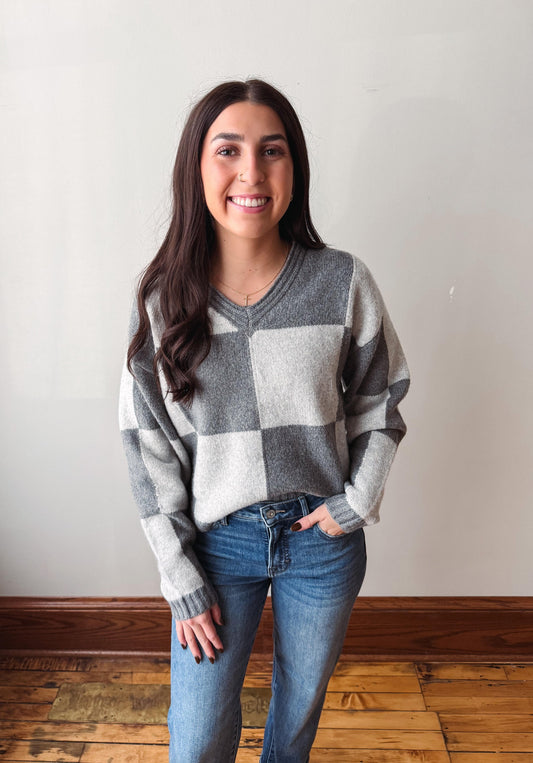 A woman with long dark hair stands indoors wearing a gray and white checkered knit sweater and blue jeans against a plain light wall and wood floor.