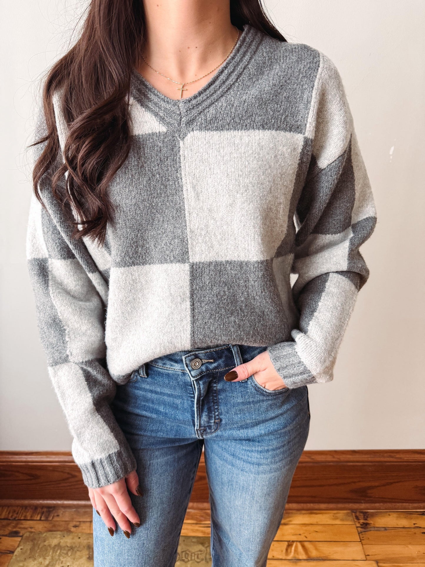 A woman wearing a gray and off-white checkered knit sweater with blue jeans, standing indoors against a light wall and wooden floor.