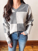 A woman wearing a gray and off-white checkered knit sweater with blue jeans, standing indoors against a light wall and wooden floor.
