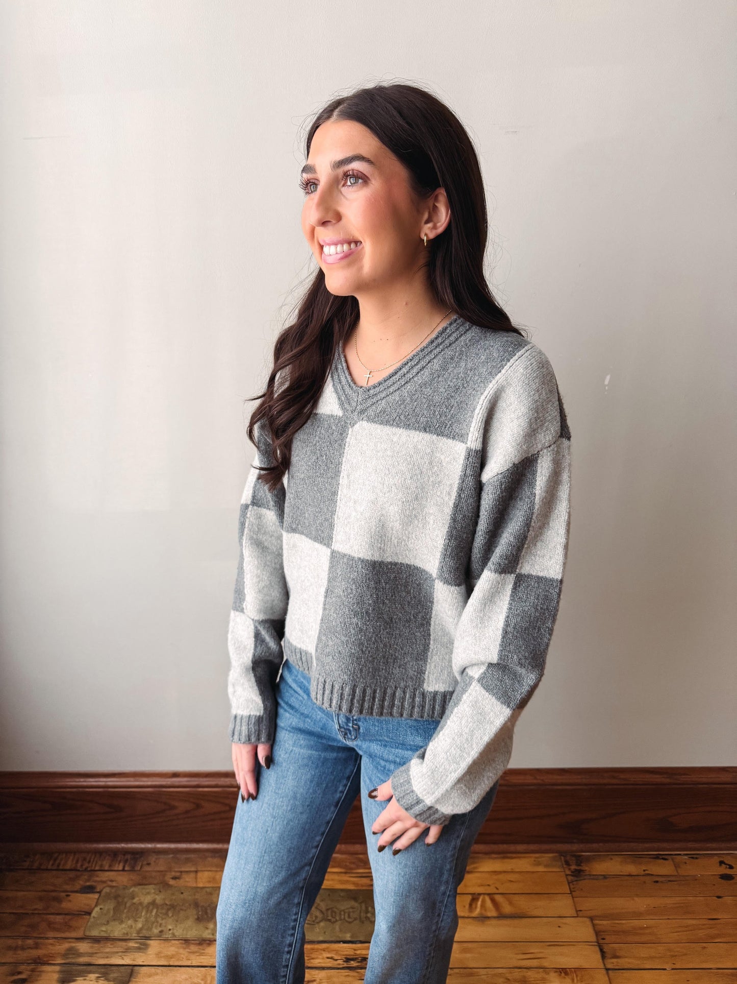 A woman wearing a gray and white checkered V-neck sweater and blue jeans, standing against a plain light gray wall with a wooden floor.