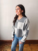 A woman wearing a gray and white checkered V-neck sweater and blue jeans, standing against a plain light gray wall with a wooden floor.