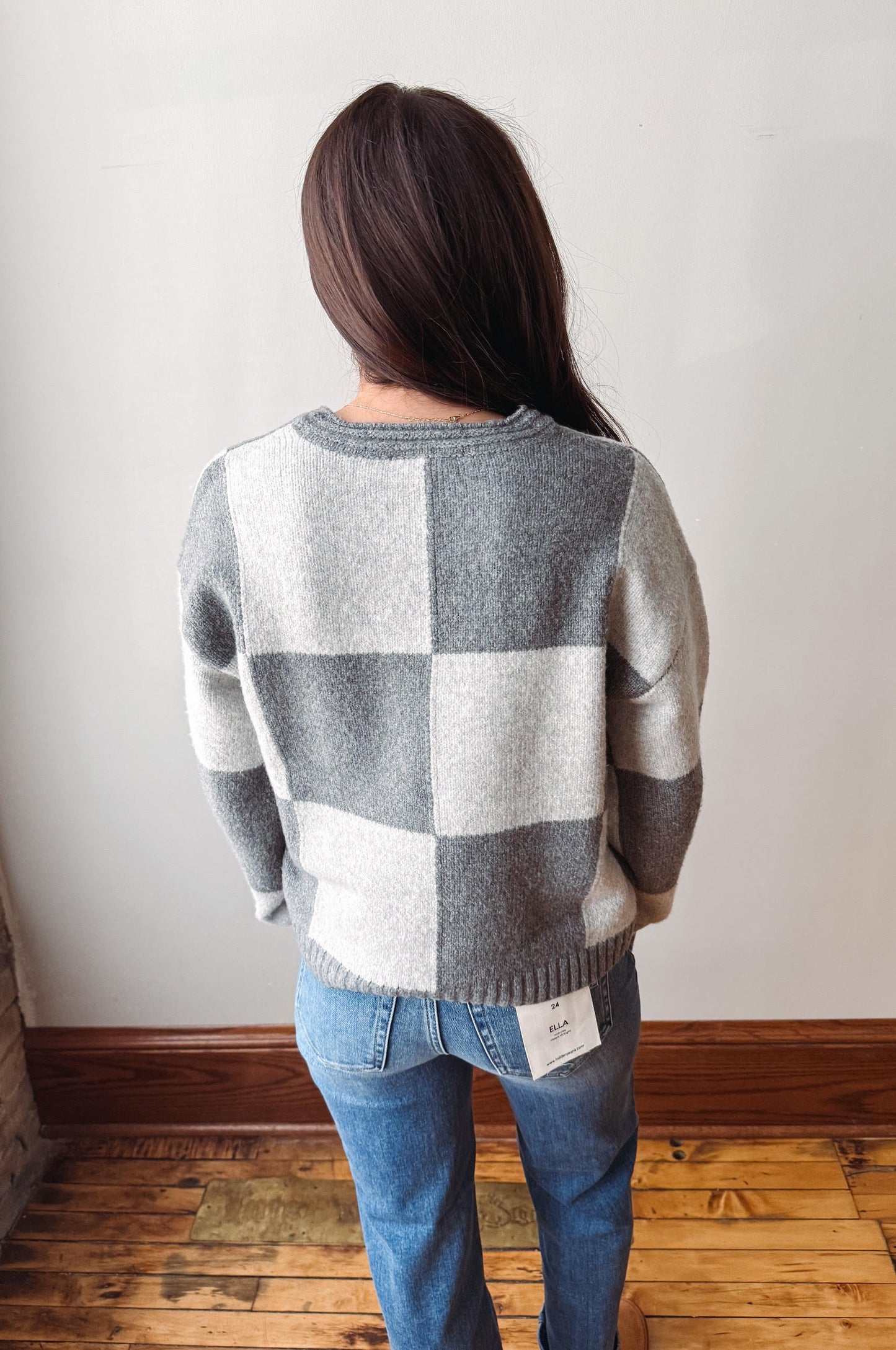 A woman viewed from behind wearing a gray and white checkered knit sweater and blue jeans, standing on a wooden floor against a plain white wall.