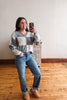 A woman takes a mirror selfie wearing a gray and white checkered V-neck sweater, blue jeans, and tan boots in a room with a wooden floor and white wall.