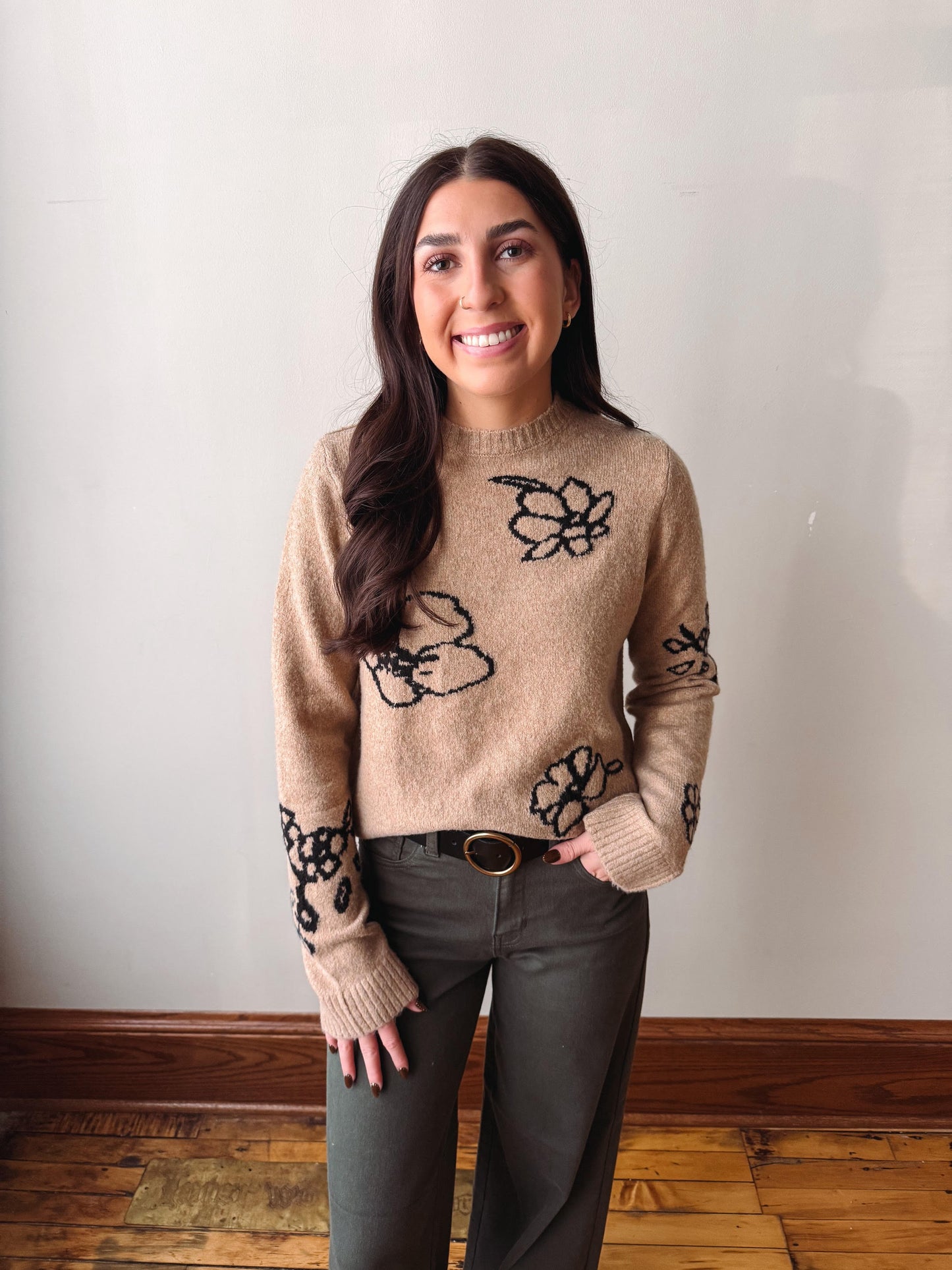 A woman with long dark hair stands indoors wearing a beige knit sweater with black floral patterns, paired with dark gray pants and a brown belt, against a plain off-white wall and wooden floor.
