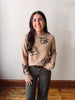 A woman with long dark hair stands indoors wearing a beige knit sweater with black floral patterns, paired with dark gray pants and a brown belt, against a plain off-white wall and wooden floor.