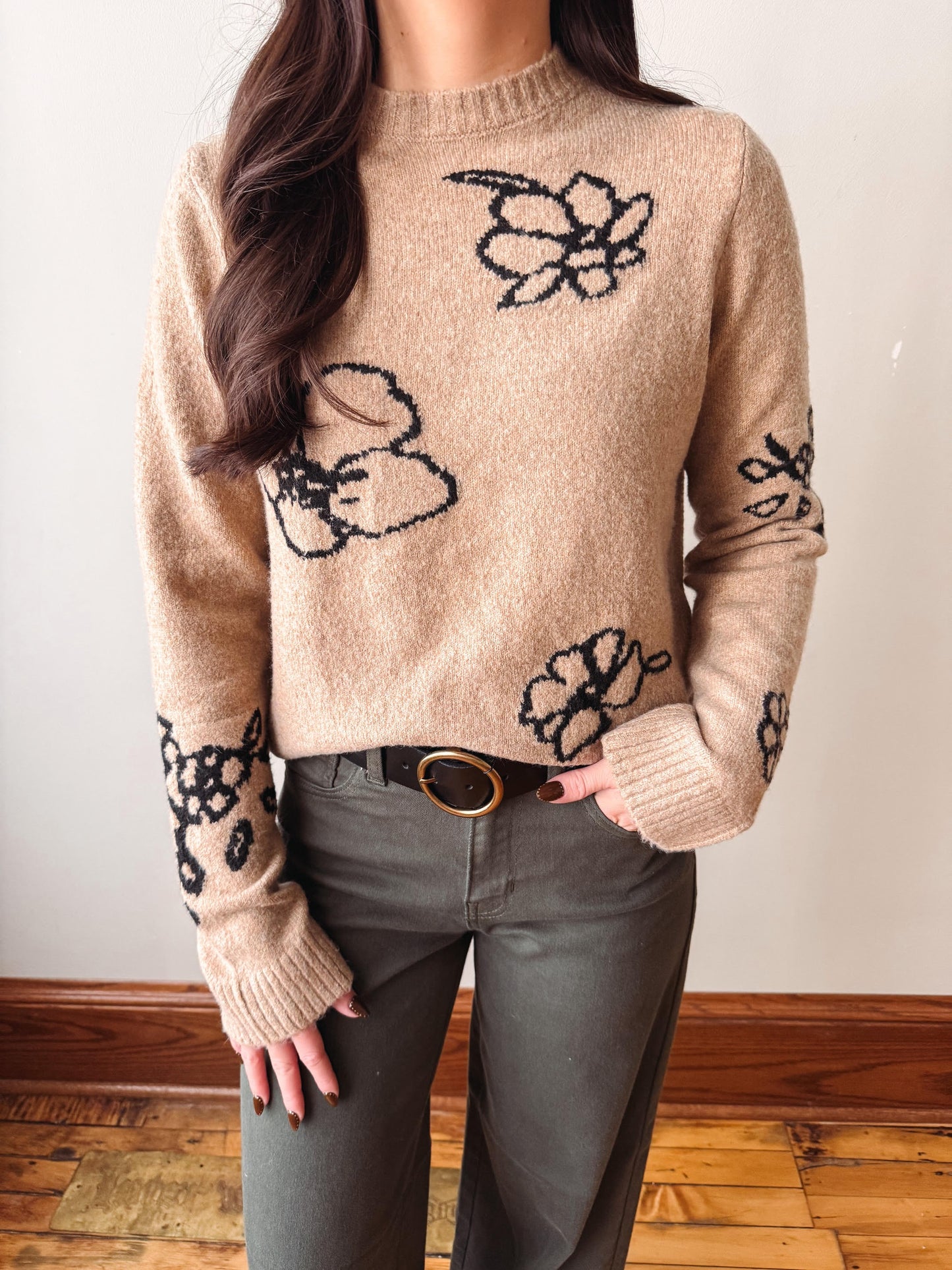 A woman wearing a beige knit sweater with black floral patterns and dark gray pants, standing indoors against a plain wall.