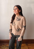 A woman wearing a beige knit sweater with black floral patterns and dark gray pants, standing against a plain light gray wall with wooden flooring visible.