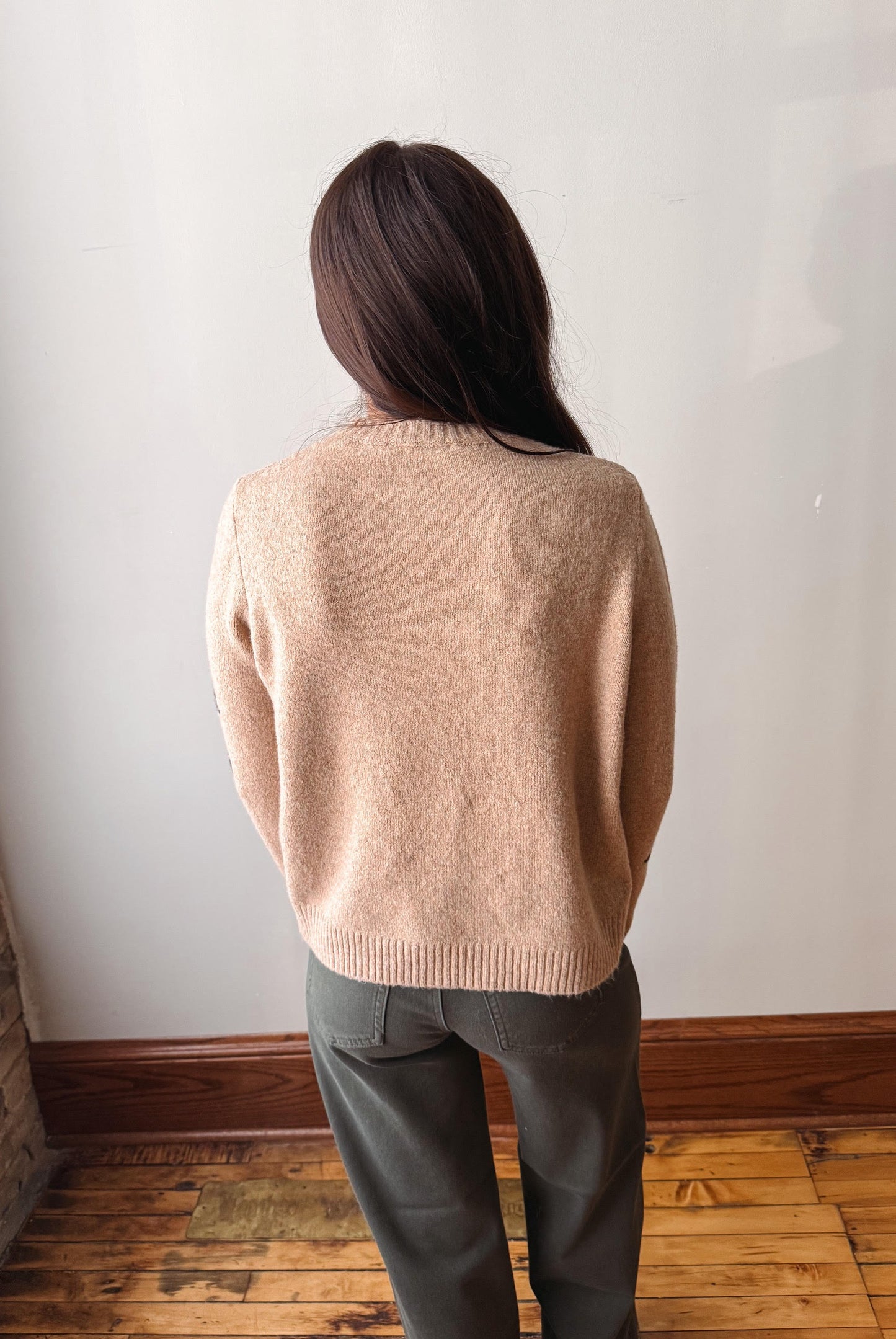 A woman viewed from behind wearing a light beige knit sweater and dark gray trousers, standing in a room with a white wall and wooden floor.