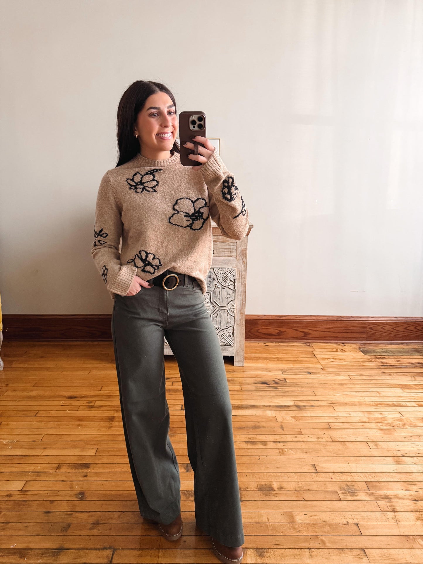 A woman takes a mirror selfie wearing a beige knit sweater with black floral patterns and dark gray wide-leg pants, standing on a wooden floor against a plain wall.