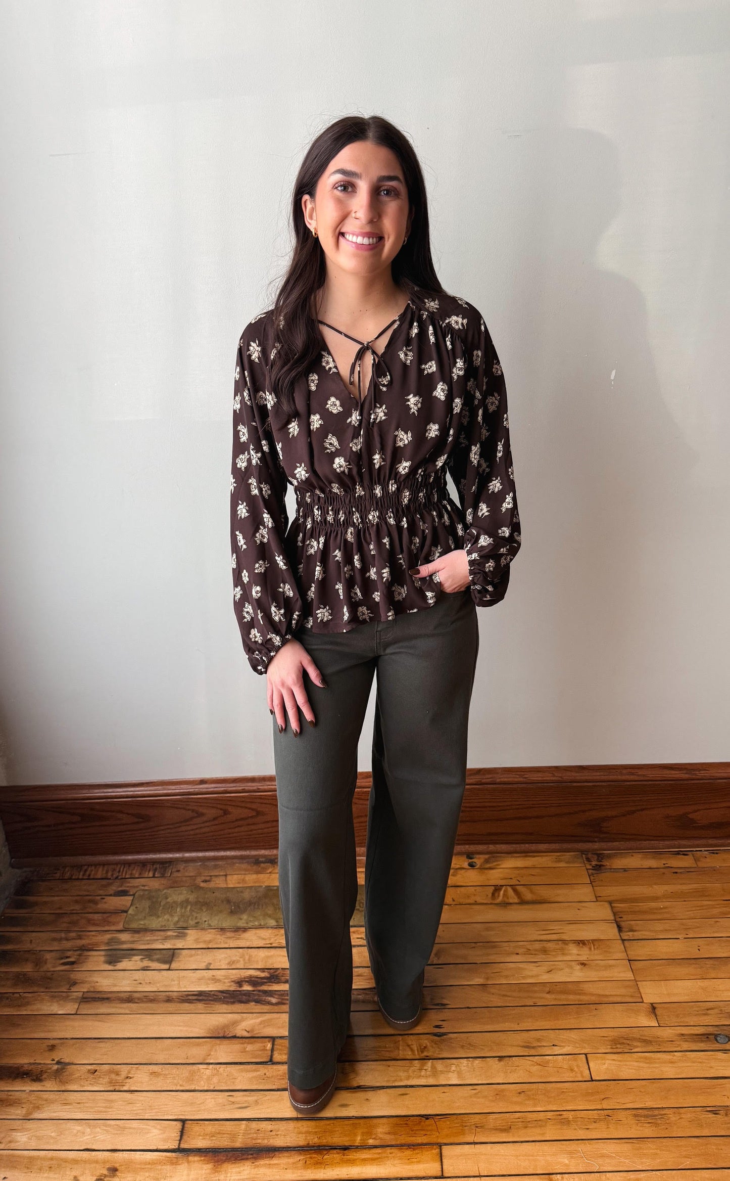 A woman stands in a room with a white wall and wooden floor, wearing a dark brown long-sleeve blouse with a white floral pattern and dark olive green wide-leg pants.