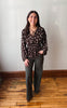 A woman stands in a room with a white wall and wooden floor, wearing a dark brown long-sleeve blouse with a white floral pattern and dark olive green wide-leg pants.