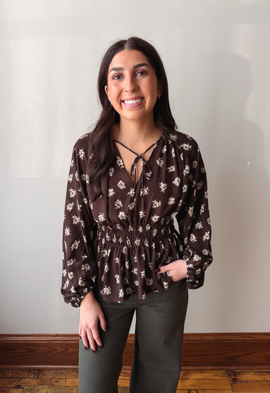 A woman wearing a brown long-sleeve blouse with a white floral pattern and a criss-cross tie neckline, paired with dark gray pants, standing against a plain off-white wall.