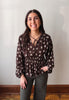 A woman wearing a brown long-sleeve blouse with a white floral pattern and a criss-cross tie neckline, paired with dark gray pants, standing against a plain off-white wall.