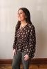A woman wearing a dark brown long-sleeve blouse with a white floral pattern and tie-neck detail, paired with dark gray pants, standing against a plain indoor wall.