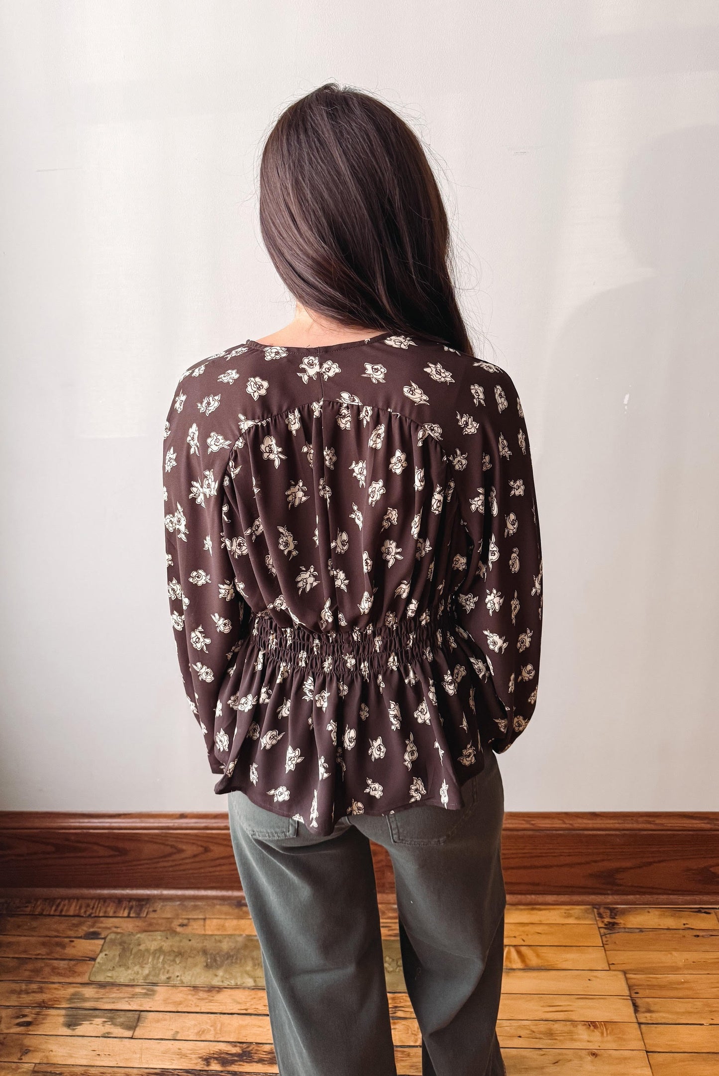 A woman viewed from behind wearing a dark brown long-sleeve blouse with a white floral pattern and a gathered smocked waist, paired with gray pants, standing in an indoor setting with a light wall and wooden floor.