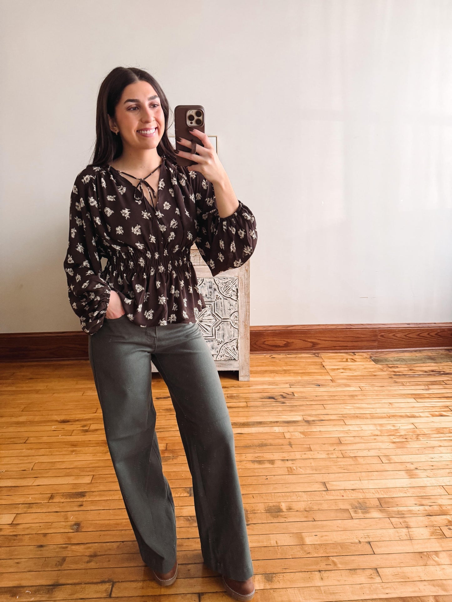 A woman takes a mirror selfie in an indoor room with hardwood floors and a white wall. She is wearing a dark brown long-sleeve blouse with a white floral pattern and dark gray wide-leg pants.