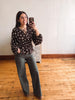 A woman takes a mirror selfie in an indoor room with hardwood floors and a white wall. She is wearing a dark brown long-sleeve blouse with a white floral pattern and dark gray wide-leg pants.