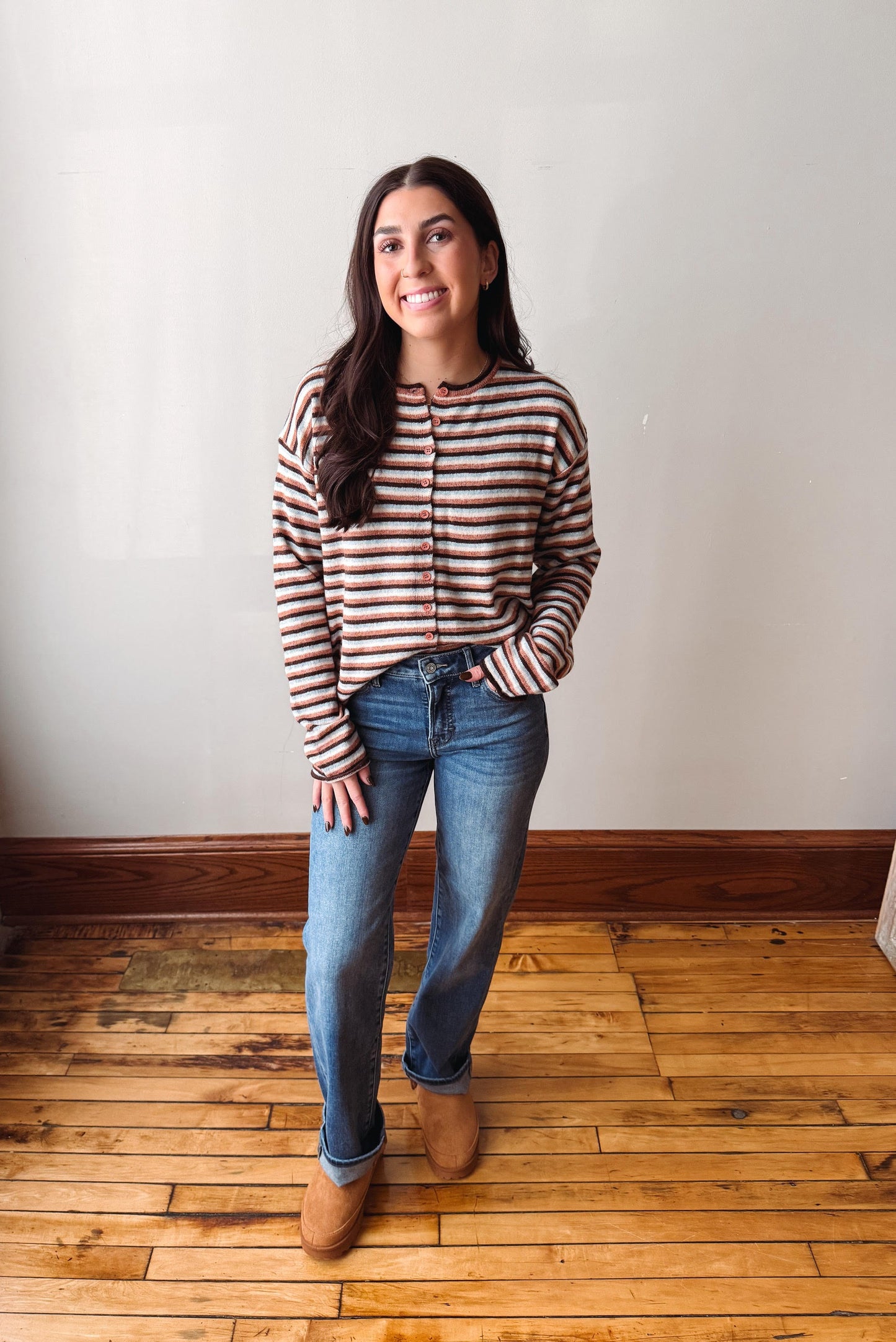 A woman stands in a room with a white wall and wooden floor, wearing a brown and beige striped long-sleeve crop top, blue jeans, and tan boots.