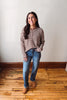 A woman stands in a room with a white wall and wooden floor, wearing a brown and beige striped long-sleeve crop top, blue jeans, and tan boots.