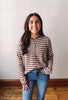 A woman with long dark hair stands indoors wearing a cropped, long-sleeve knit top with horizontal stripes in brown, beige, and white, paired with blue jeans.