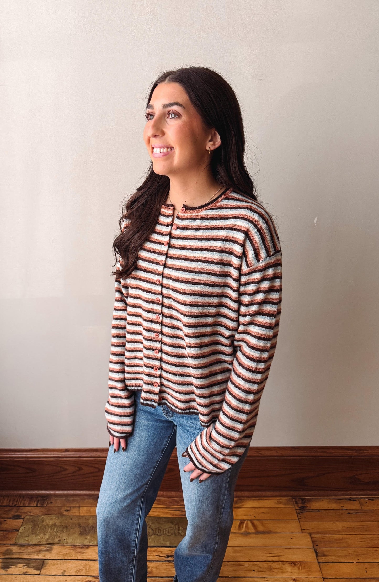 A woman with long dark hair stands in a room wearing a long-sleeve knit top with horizontal stripes in brown, beige, and white, paired with blue jeans.