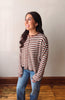 A woman with long dark hair stands in a room wearing a long-sleeve knit top with horizontal stripes in brown, beige, and white, paired with blue jeans.