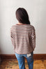 A woman viewed from behind wearing a long-sleeve knit sweater with thin horizontal stripes in rust orange, brown, and cream, paired with blue jeans, standing against a plain off-white wall.