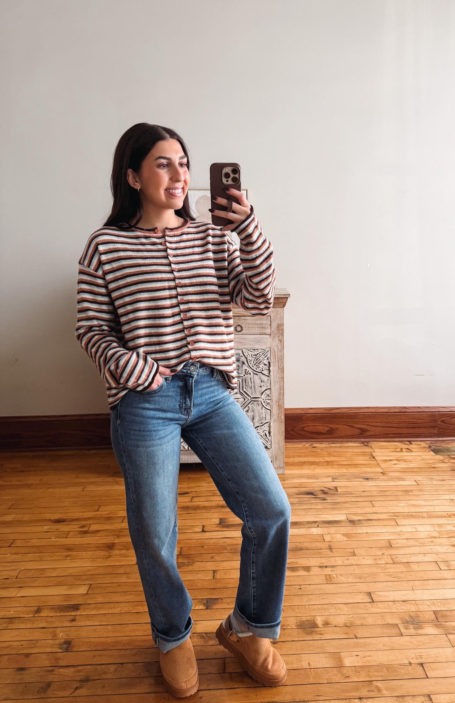 A woman takes a mirror selfie wearing a brown and white striped long-sleeve knit top, blue jeans, and tan suede boots, standing in a room with a wooden floor and white wall.