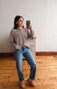 A woman takes a mirror selfie wearing a brown and white striped long-sleeve knit top, blue jeans, and tan suede boots, standing in a room with a wooden floor and white wall.