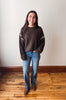 A woman stands in a room with a light-colored wall and wooden floor, wearing a dark brown knit sweater with white sleeve stripes, blue jeans, and brown boots.