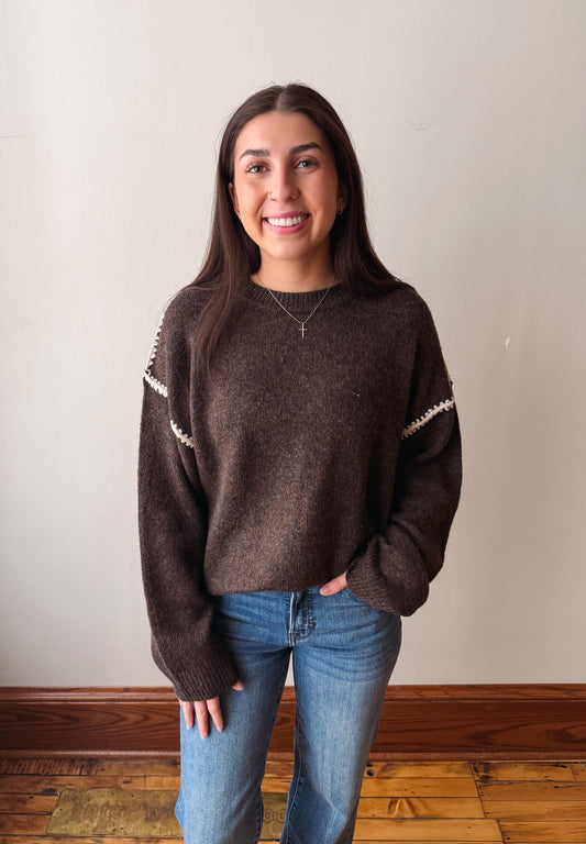A woman with long dark hair stands indoors wearing a brown knit sweater with white patterned trim on the shoulders and blue jeans, against a plain light wall with a wooden baseboard and floor.