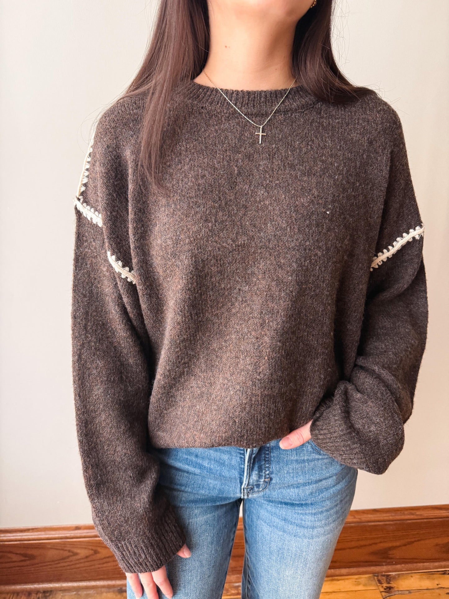 A person wearing a brown knit sweater with white decorative trim along the shoulders and sleeves, paired with blue jeans, standing indoors against a light wall.