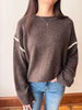 A person wearing a brown knit sweater with white decorative trim along the shoulders and sleeves, paired with blue jeans, standing indoors against a light wall.