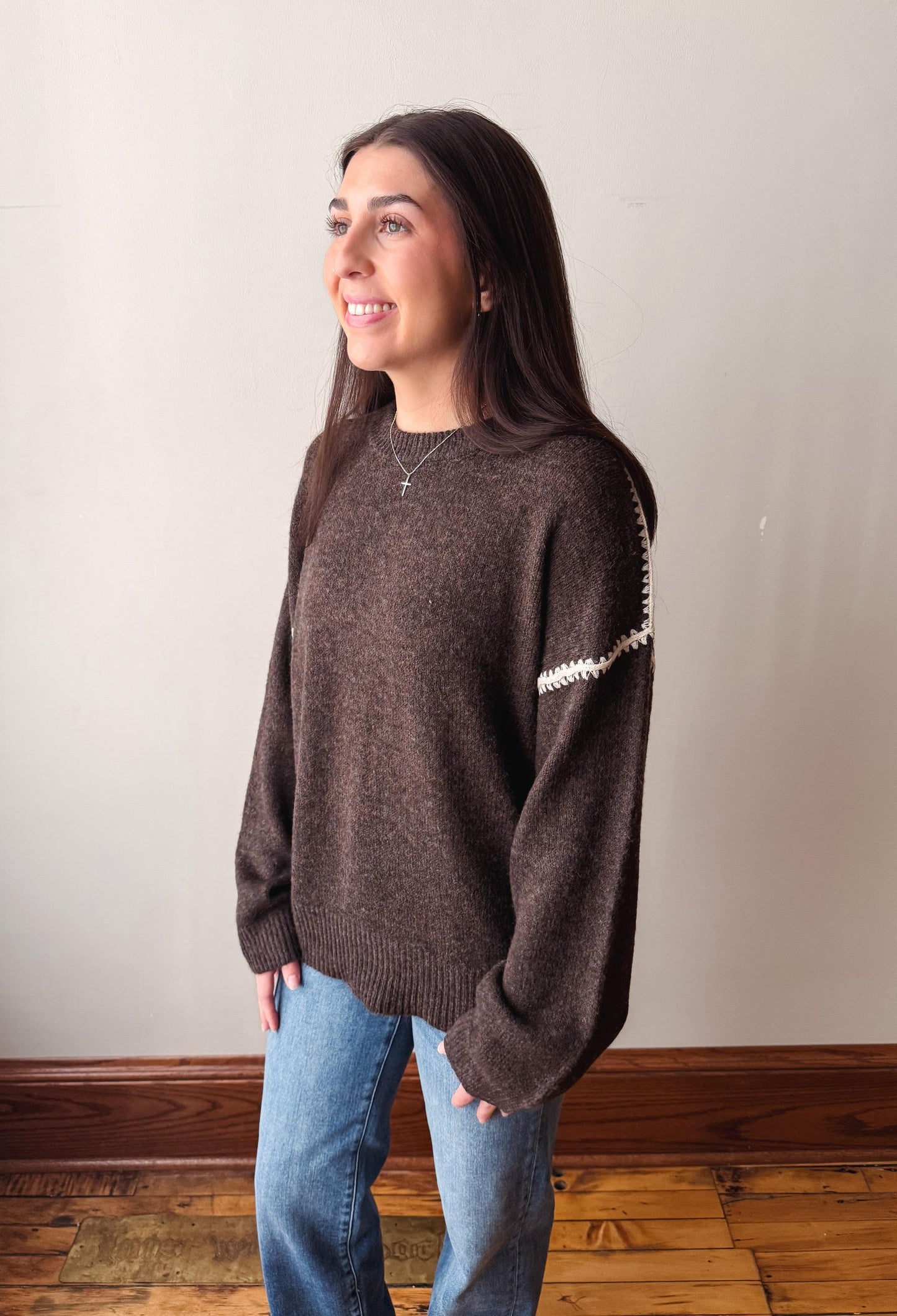 A woman wearing a dark brown knit sweater with white textured trim along the shoulder, paired with blue jeans, standing in a room with a light beige wall and wooden floor.