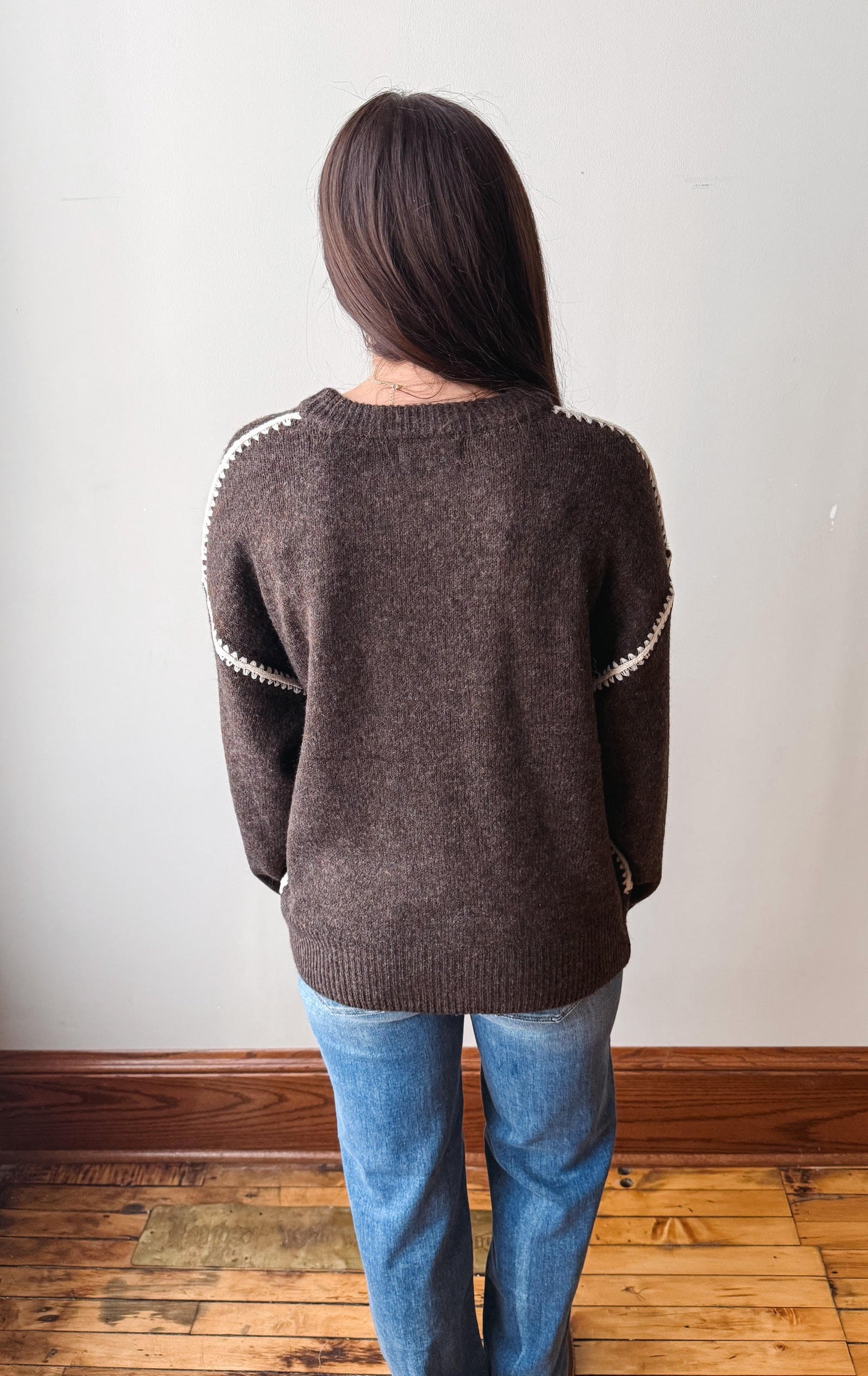 A person viewed from behind wearing a dark brown knit sweater with white trim along the shoulders and sleeves, paired with blue jeans, standing on a wooden floor against a plain light wall.