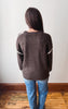 A person viewed from behind wearing a dark brown knit sweater with white trim along the shoulders and sleeves, paired with blue jeans, standing on a wooden floor against a plain light wall.