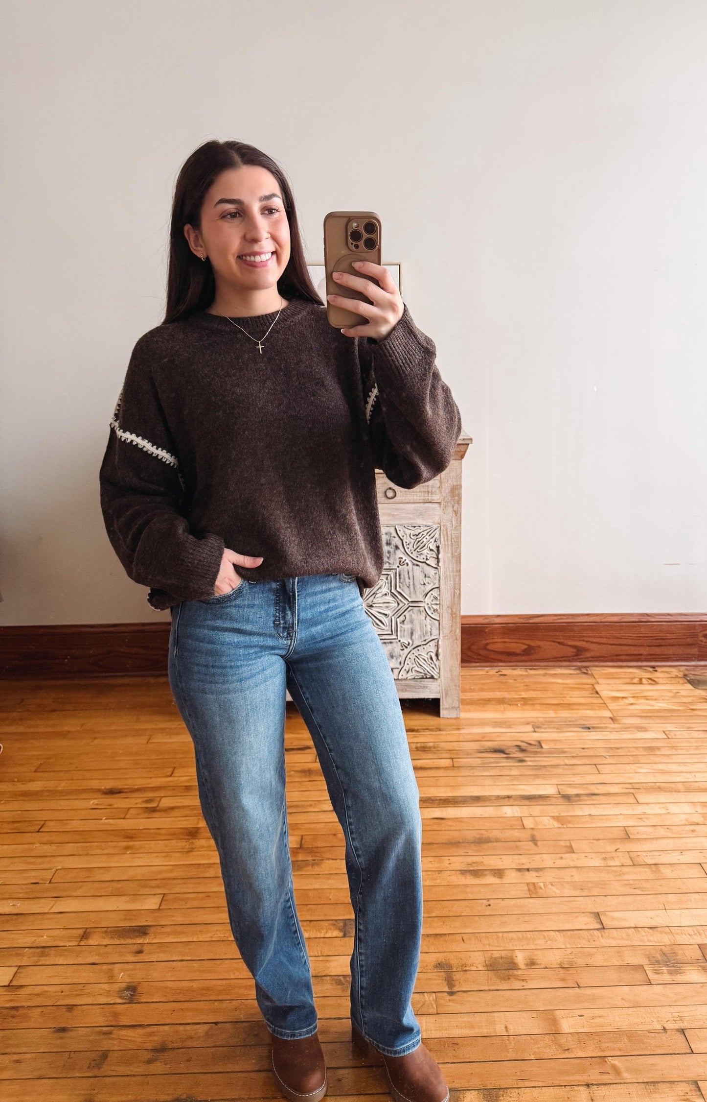 A woman takes a mirror selfie wearing a dark brown knit sweater with white sleeve stripes, blue jeans, and brown boots in a room with a wooden floor and white wall.