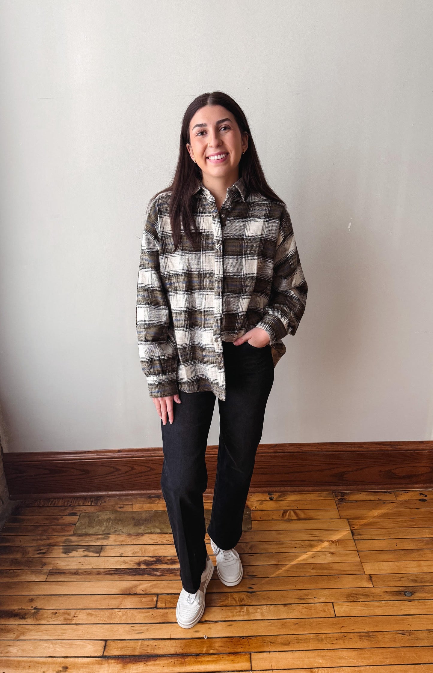 A woman stands in a room with a white wall and wooden floor, wearing a black, white, and gray plaid button-down shirt, black pants, and white sneakers.