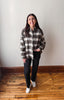 A woman stands in a room with a white wall and wooden floor, wearing a black, white, and gray plaid button-down shirt, black pants, and white sneakers.