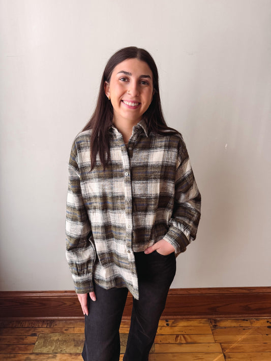 A woman with long dark hair stands indoors wearing a brown, beige, and white plaid button-up shirt and black pants, with her hands in her pockets, against a plain off-white wall and wooden floor.