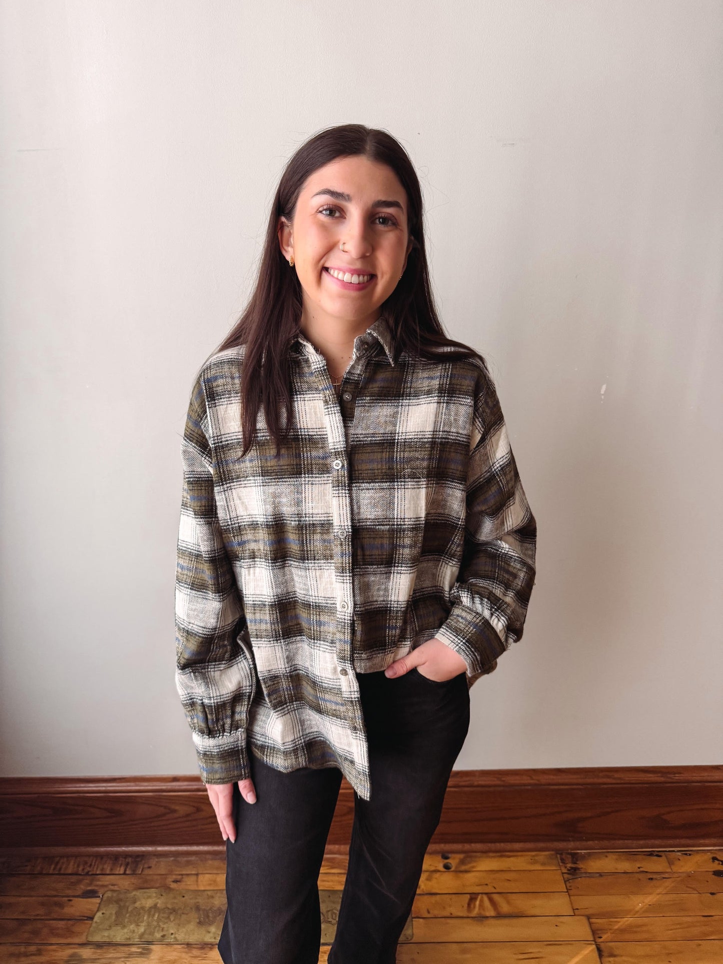 A woman with long dark hair stands indoors wearing a brown, beige, and white plaid button-up shirt and black pants, with her hands in her pockets, against a plain off-white wall and wooden floor.