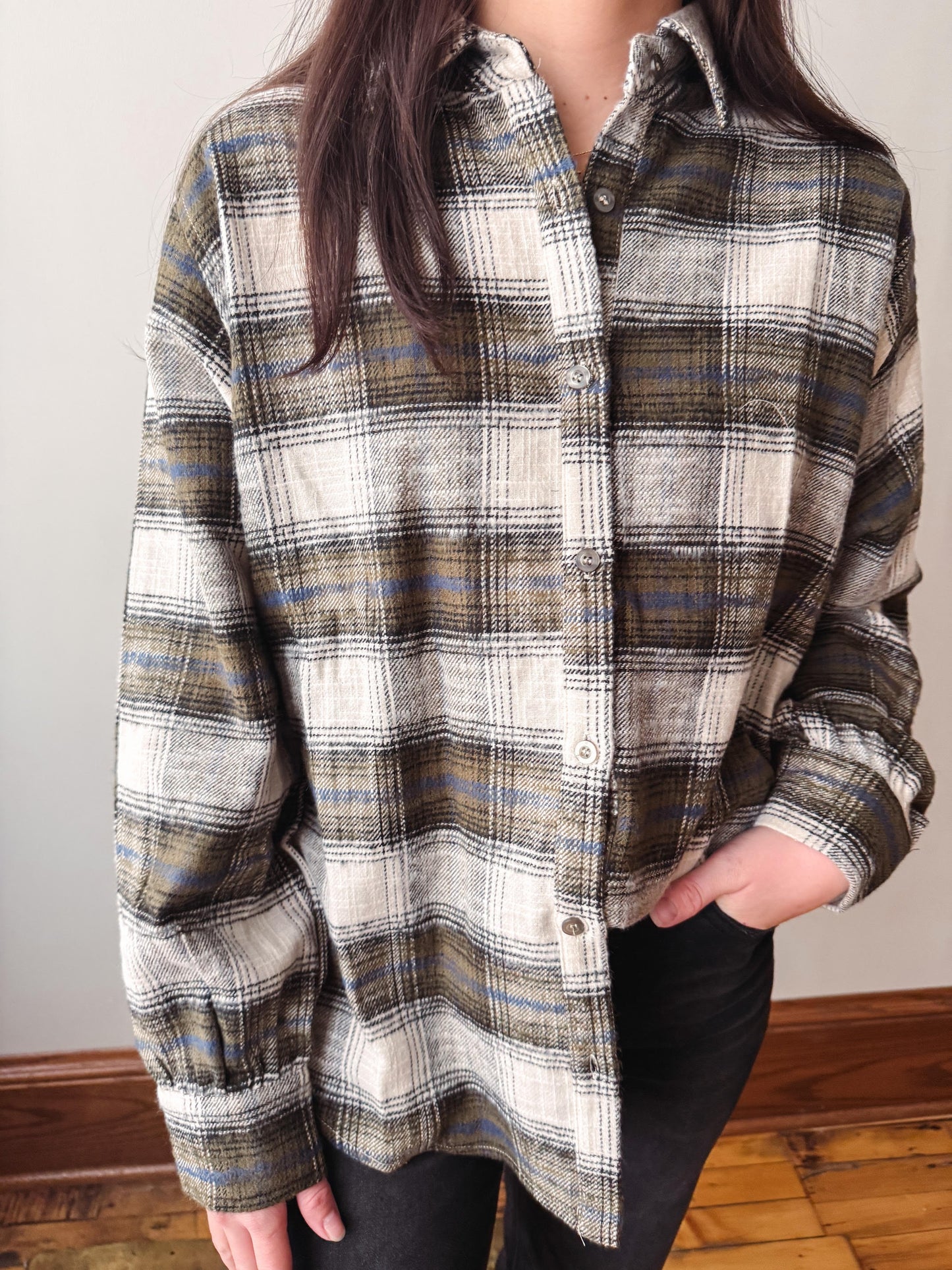 A person wearing an oversized long-sleeve plaid button-up shirt in cream, brown, olive green, and blue tones, paired with black pants, standing indoors against a plain wall.