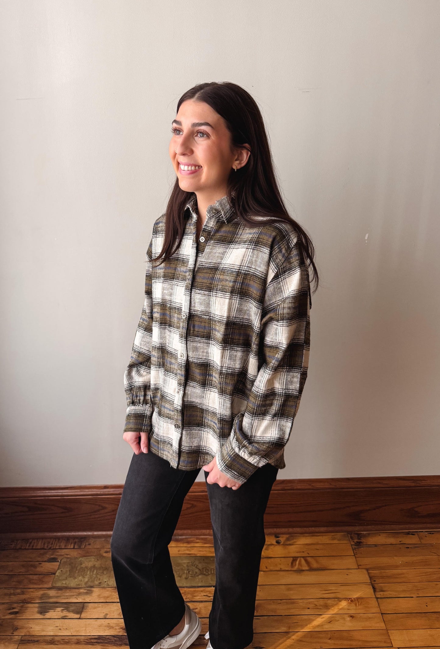 A woman modeling a long-sleeve plaid button-up shirt in brown, beige, and white, paired with black pants, standing in a room with a light gray wall and wooden floor.